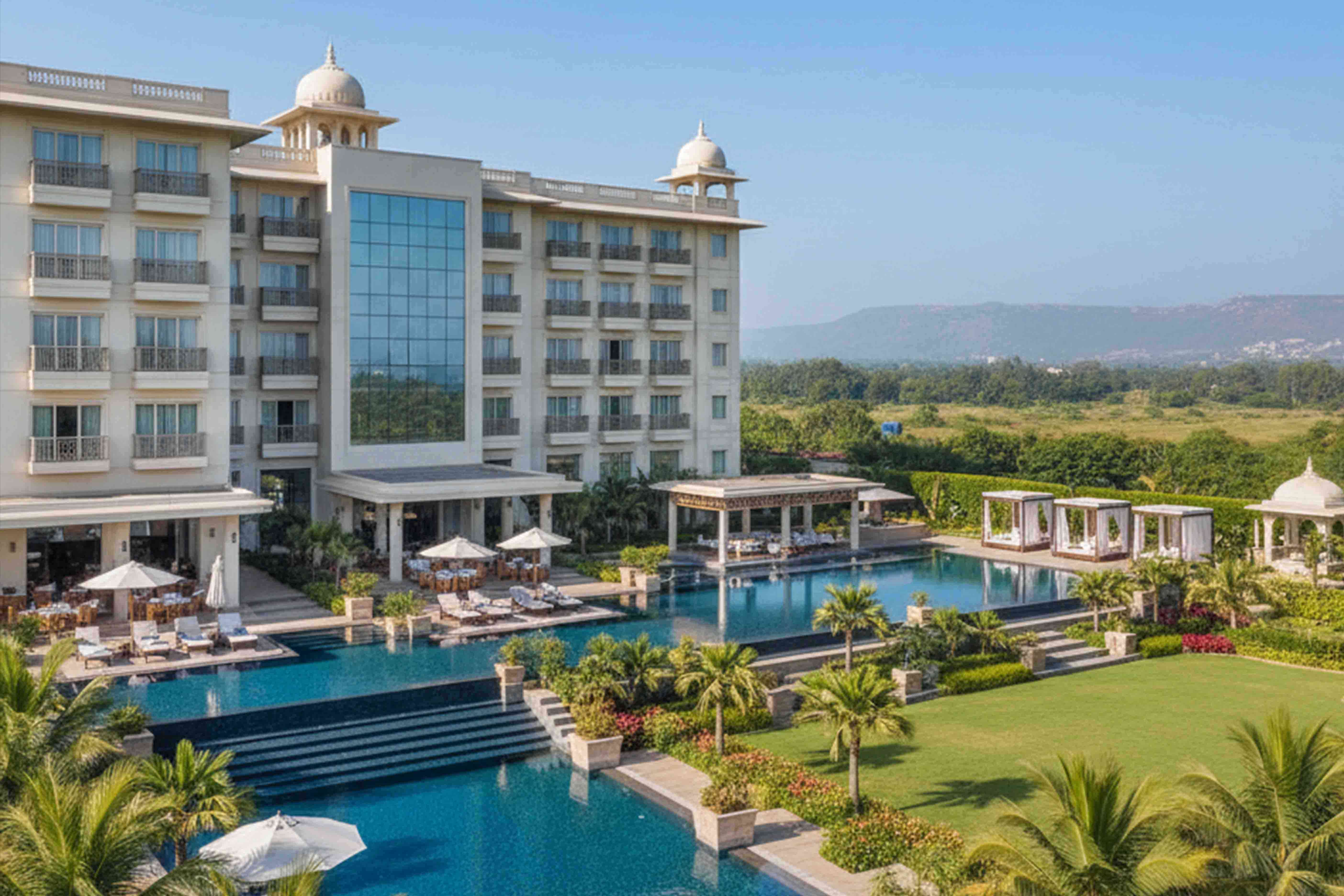 Luxury Indian hotel with large pool, green lawns, and hills in the background.